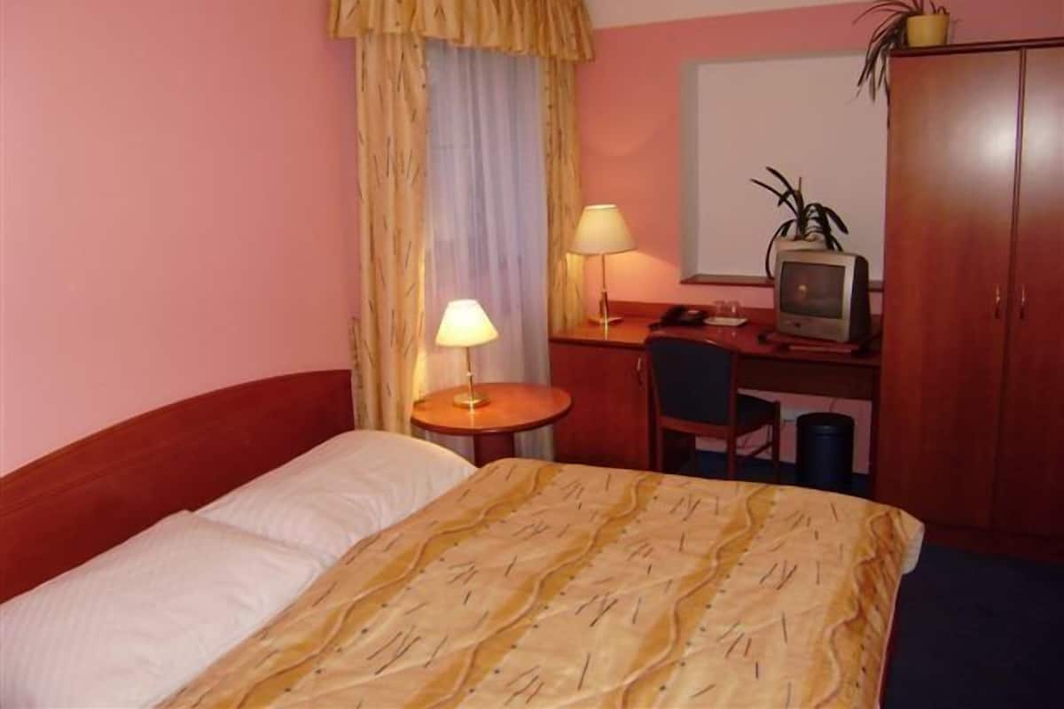 A hotel room with a bed, a desk with a computer, a chair, a lamp, a window with curtains, and a wardrobe.
