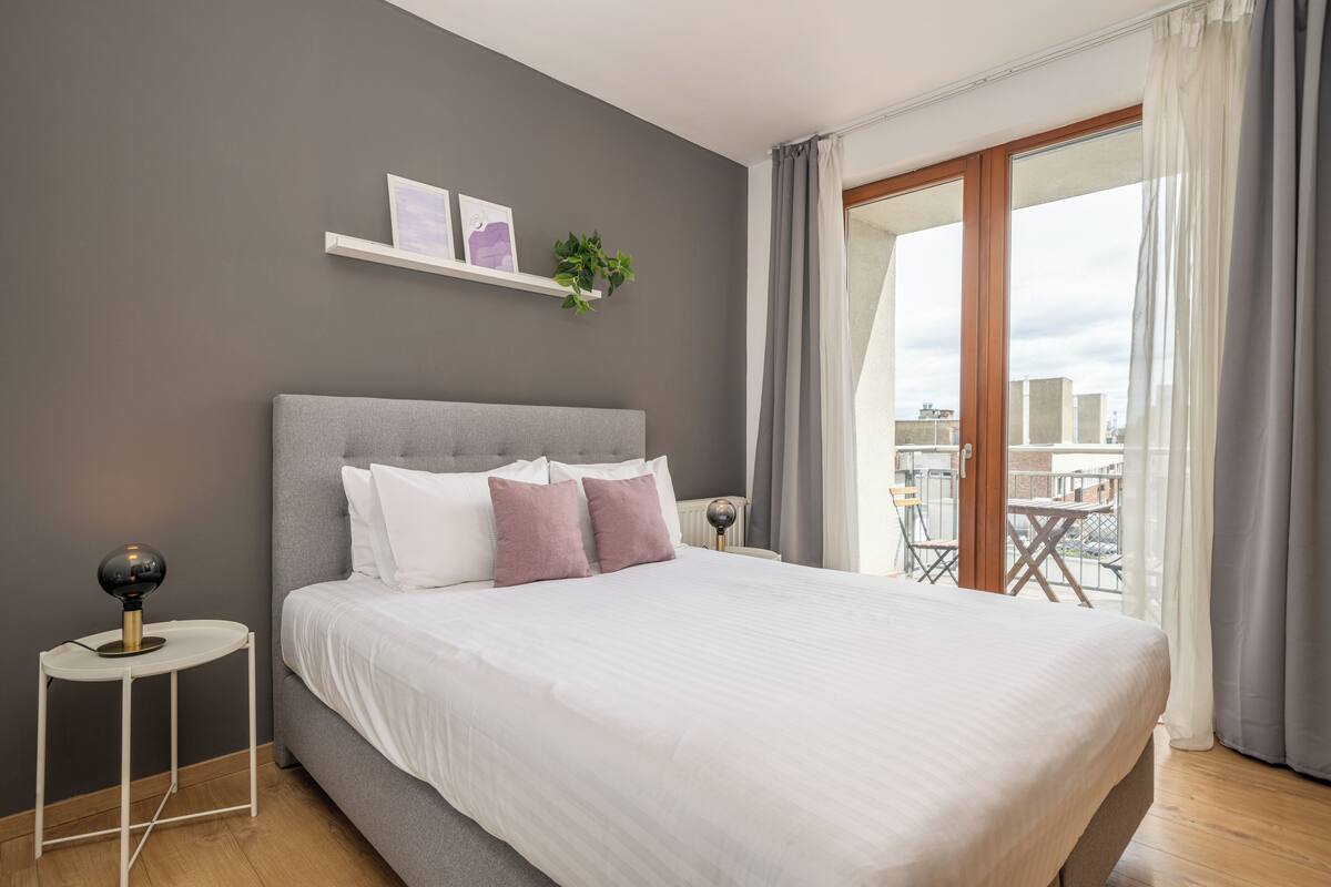 Une chambre moderne avec un grand lit, une table de chevet, une petite étagère avec des photos et une plante, et une vue sur la ville par une grande fenêtre.