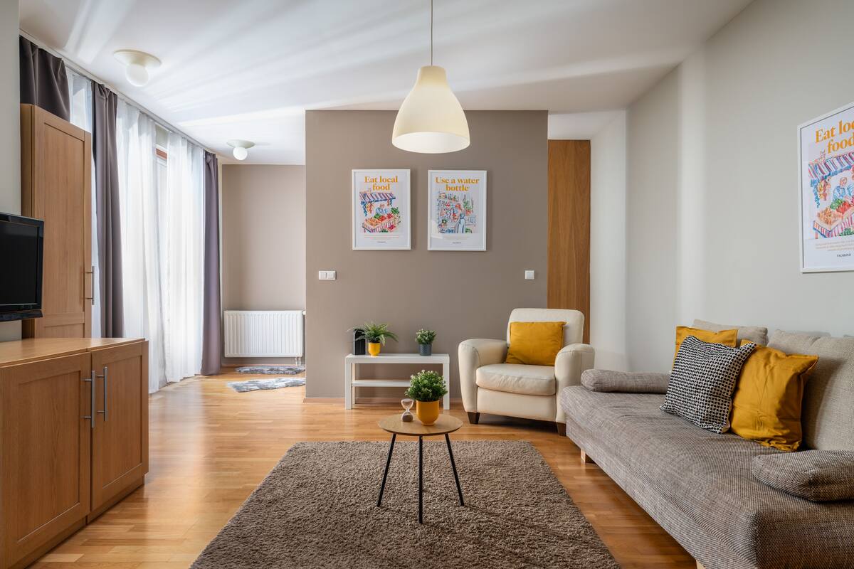 Un salon moderne avec un canapé, une table basse et un téléviseur. Deux affiches encadrées ornent le mur, et un luminaire est suspendu au plafond.