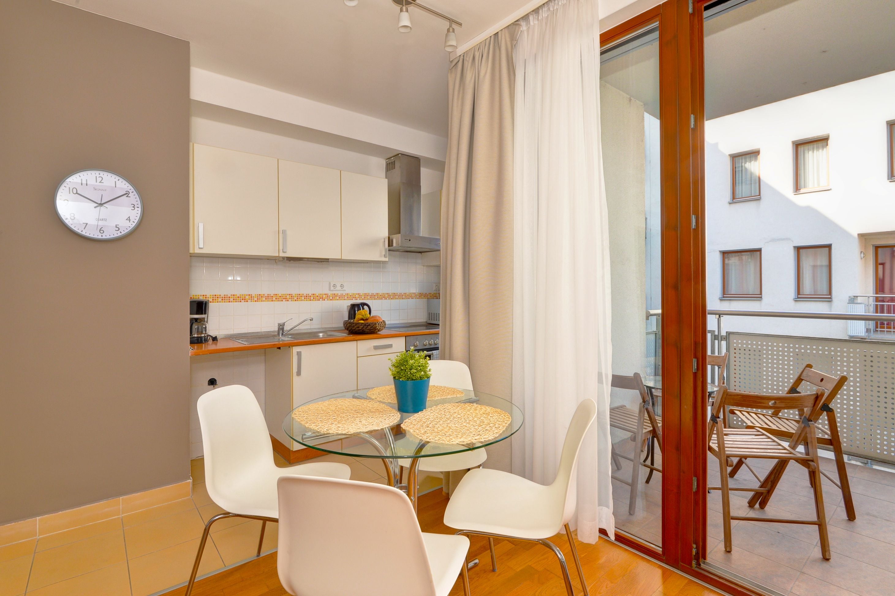 Cozinha moderna com armários brancos, um relógio e um espaço de refeições com mesa de vidro e cadeiras brancas.