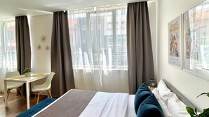 Habitación de hotel con una cama, un escritorio y una silla. Hay cortinas en las ventanas y una planta sobre la mesita de noche.