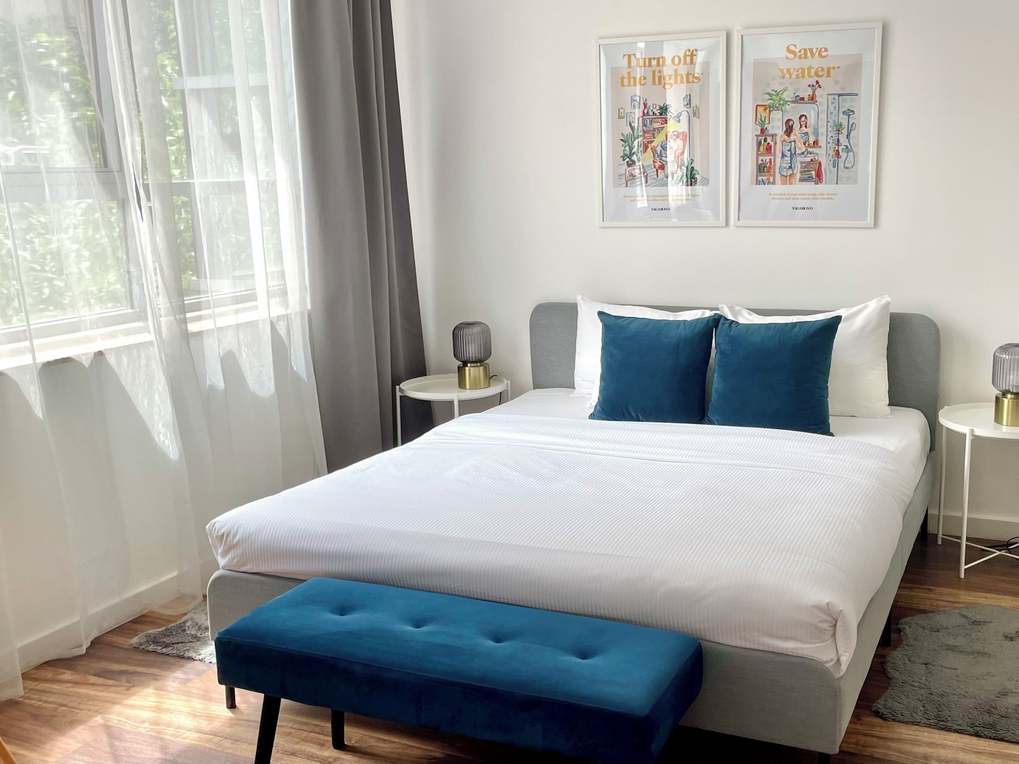 A modern bedroom with a large bed, bedside tables, a bench, and two framed posters on the wall.