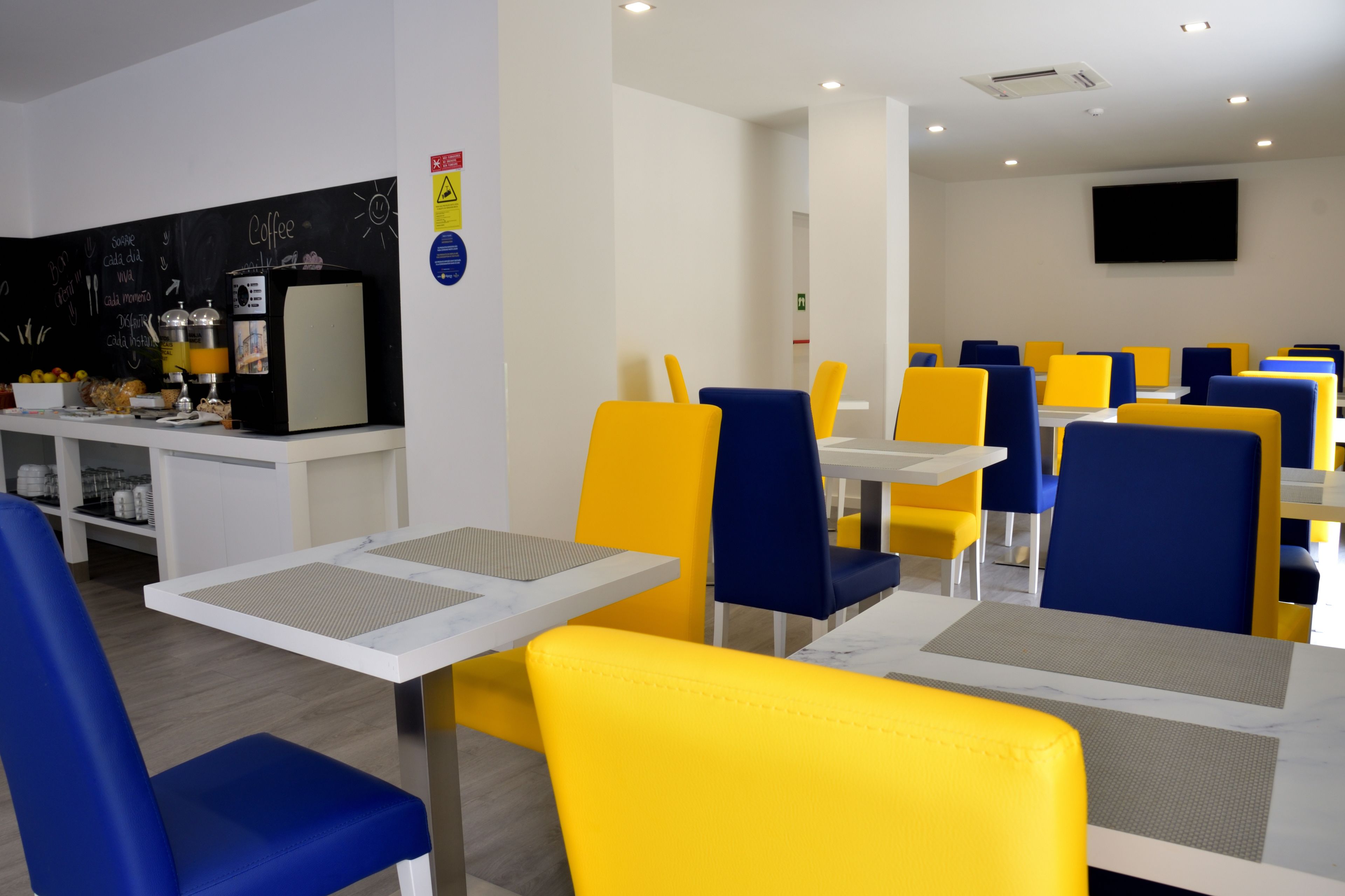 A dining area with yellow and blue chairs, a coffee station, and a flat-screen TV on the wall.