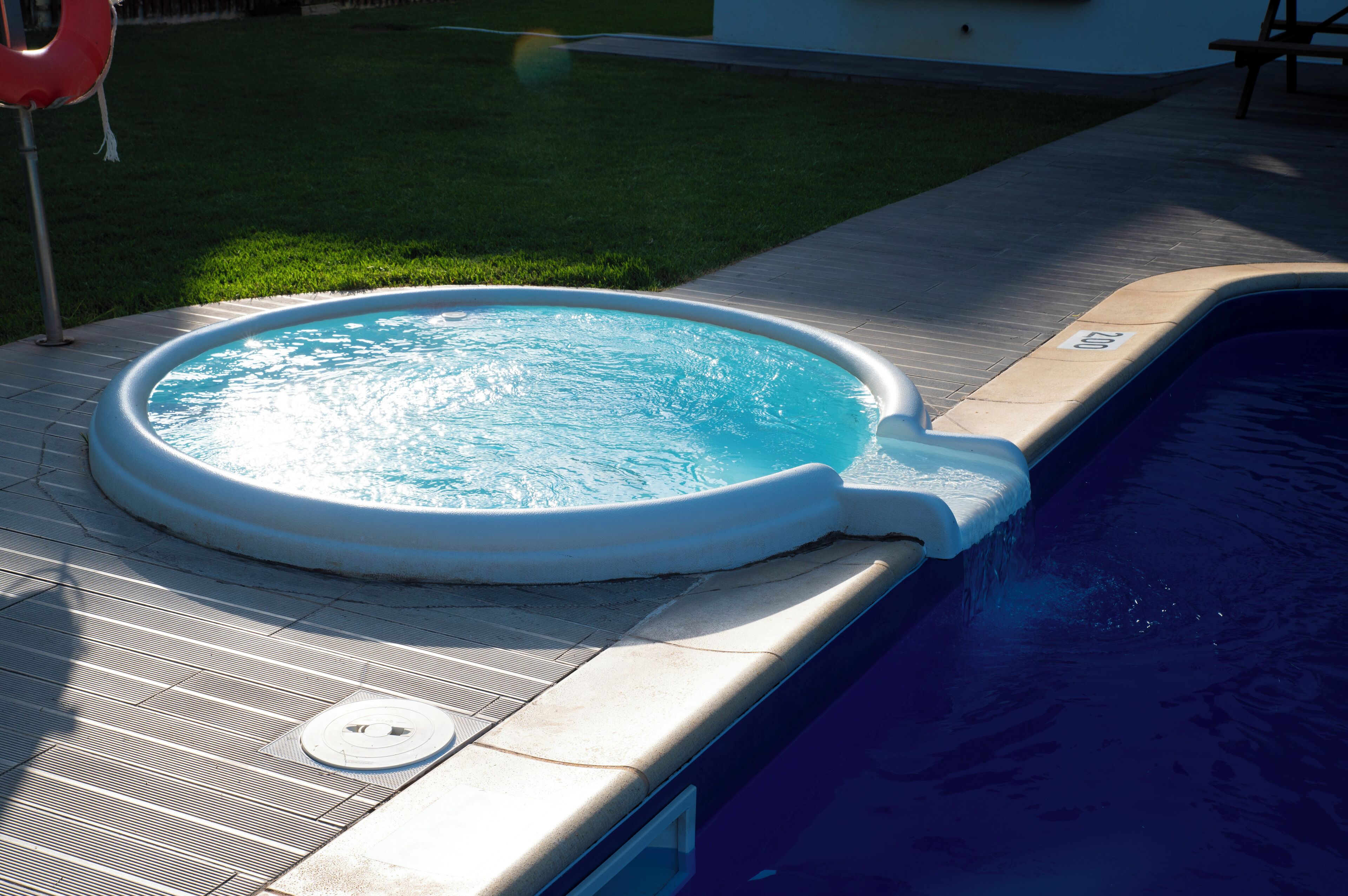 Een zwembad met een jacuzzi, omgeven door een betegeld zonnedek en een gazon.