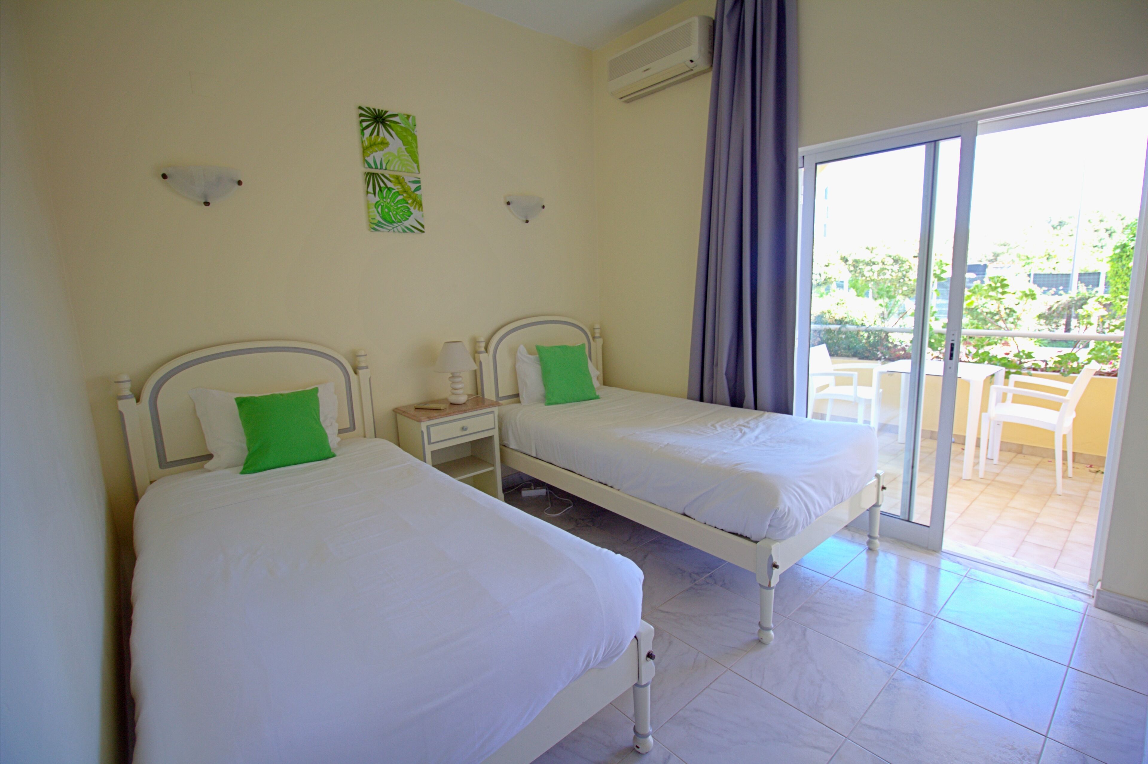 Une chambre d’hôtel avec deux lits simples, une petite table, une table de chevet et une porte-fenêtre coulissante donnant sur un espace extérieur aménagé avec des chaises.