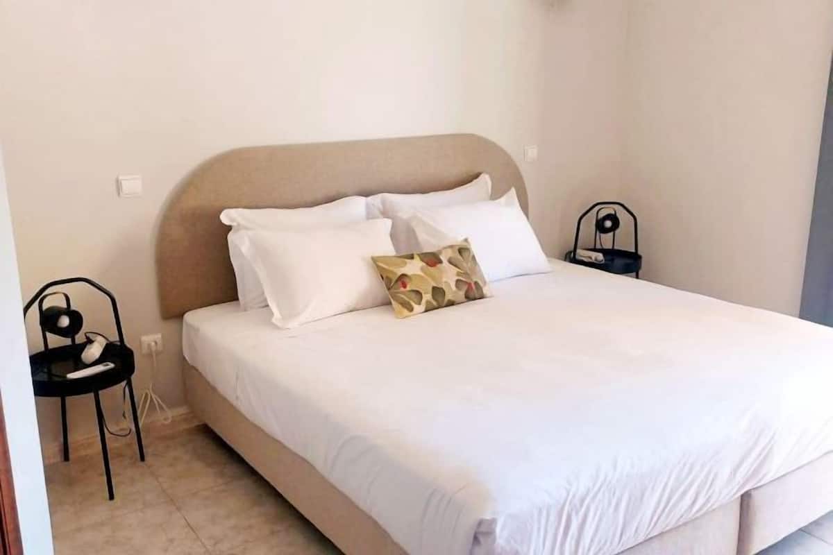 A neatly made bed with white bedding and a patterned pillow, a small bedside table with a lamp, and a chair with a phone and charger.