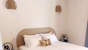 Ein ordentlich bezogenes Bett mit weißen Laken und einem einzelnen Zierkissen, ein kleiner Nachttisch mit Lampe sowie eine an der Wand montierte Klimaanlage.