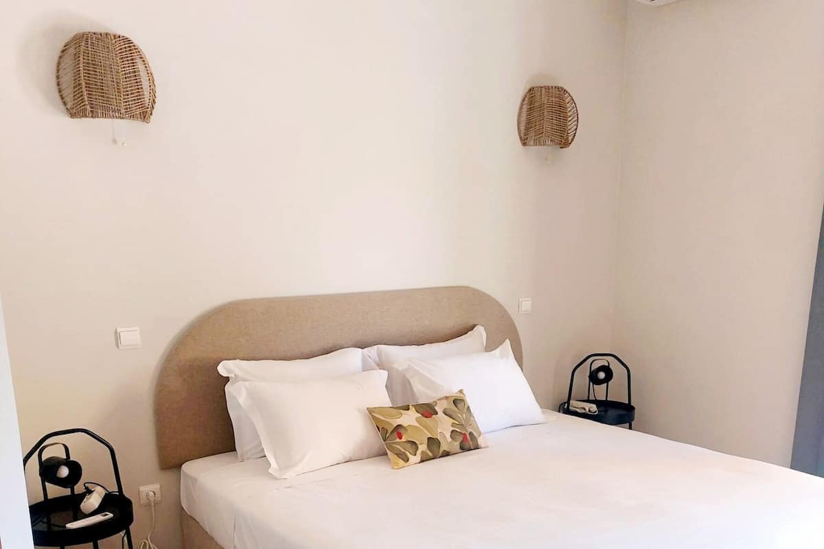 A neatly made bed with white linens and a single decorative pillow, a small bedside table with a lamp, and an air conditioning unit mounted on the wall.