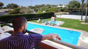 Un homme se tient au bord d’une piscine, avec des chaises longues et un petit bâtiment à proximité.