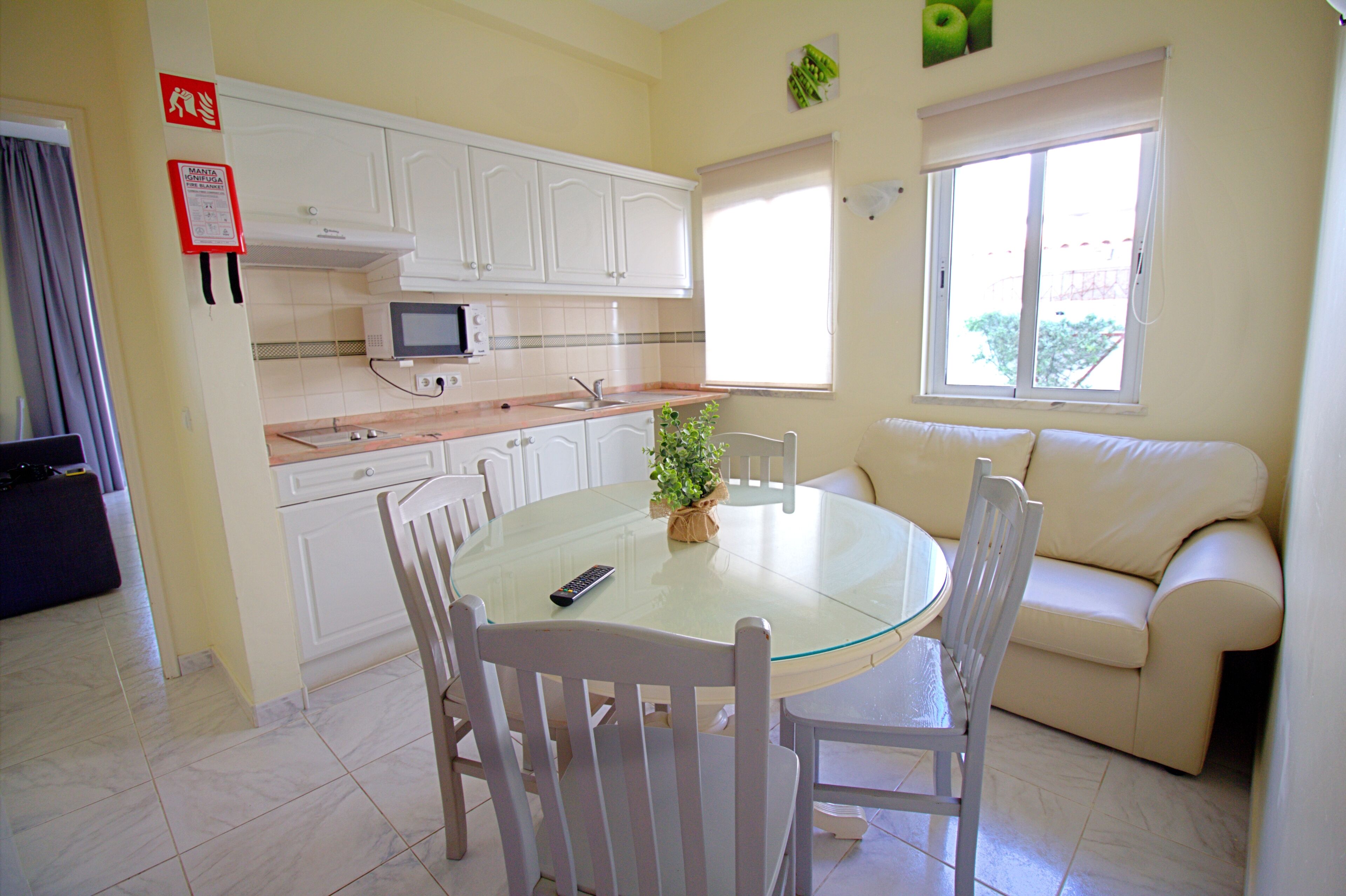 Cocina compacta con armarios blancos, microondas y comedor con mesa de vidrio y sillas blancas.