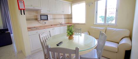 Een kleine keuken met witte kastjes, een magnetron, en een eethoek met een glazen tafel en witte stoelen.