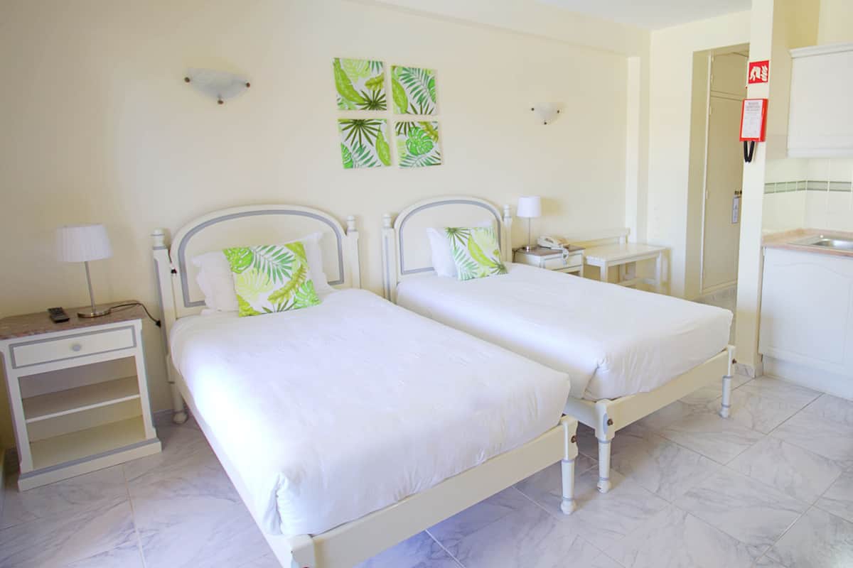 A hotel room with two single beds, a nightstand with a lamp, a small table, and a wall-mounted telephone.