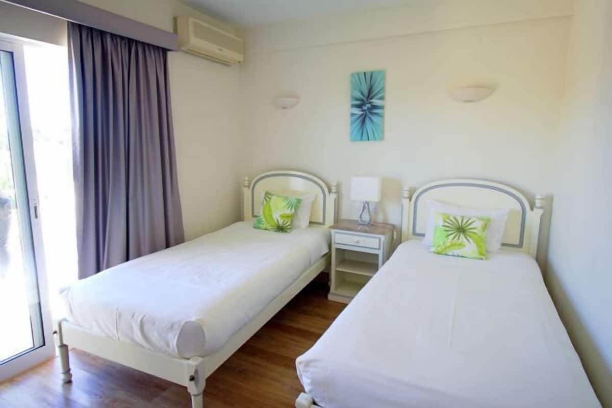 A room with two single beds, a nightstand, a lamp, a window with curtains, and a wall-mounted light fixture.