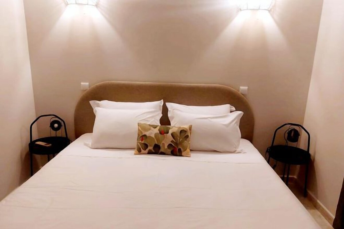 A neatly made bed with white linens and two pillows, a patterned throw pillow, and two wall-mounted reading lamps.