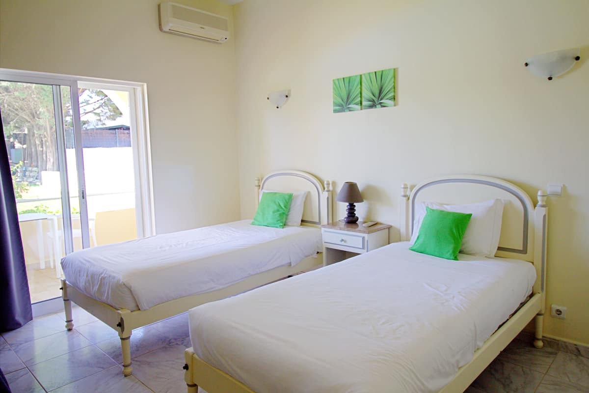 A room with two beds, each with white bedding and a green pillow. There is a nightstand with a lamp between the beds.