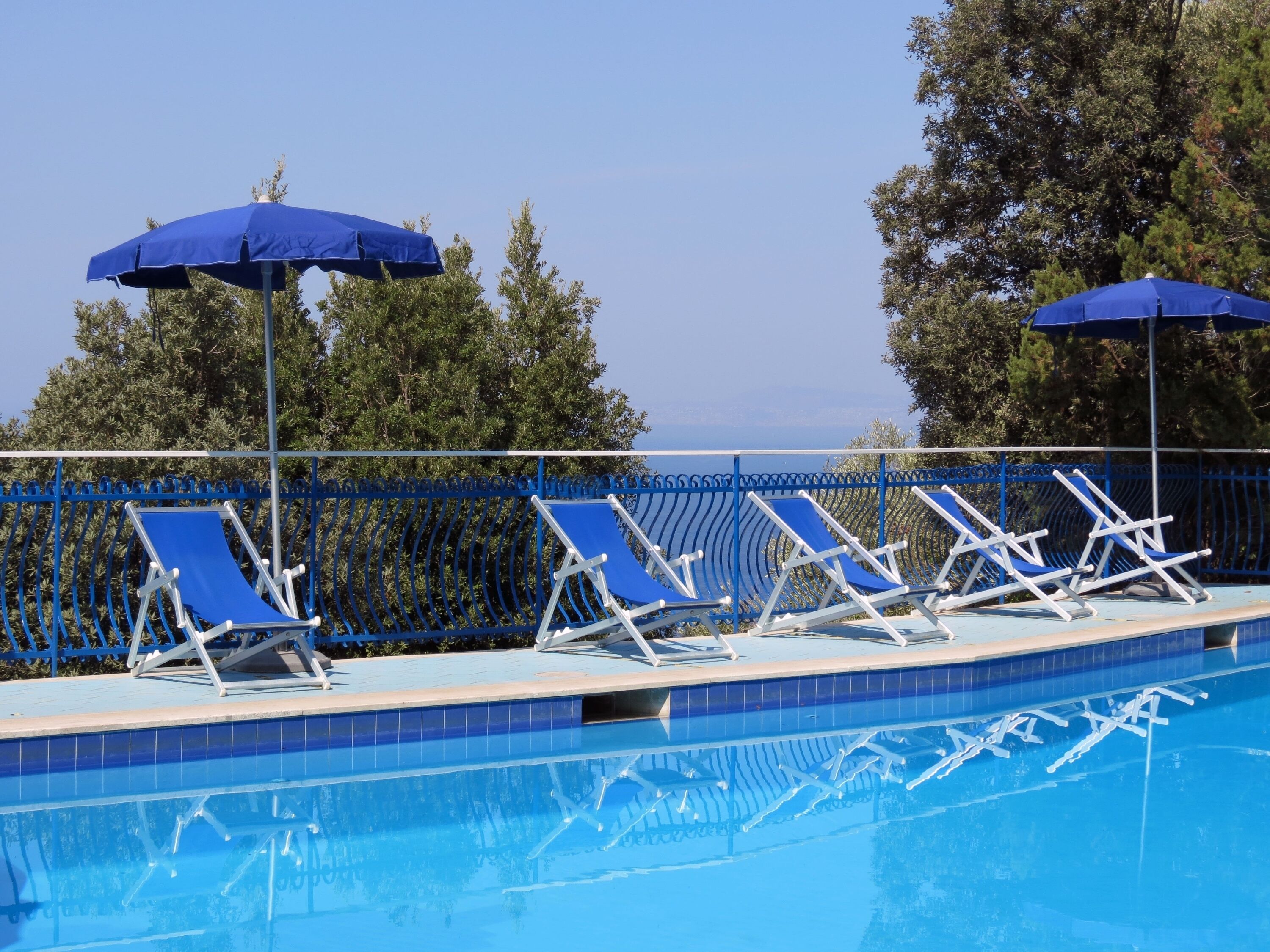 A swimming pool with blue lounge chairs and umbrellas, surrounded by a blue fence and trees.