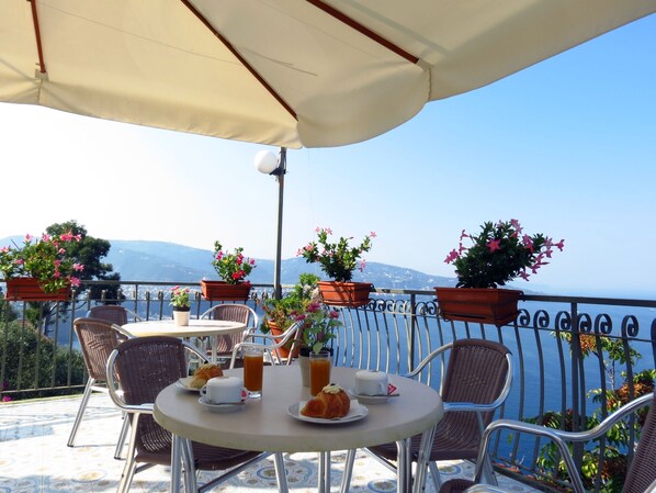 A terrace with round tables and wicker chairs, potted plants, and a view of the sea and mountains.