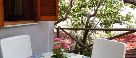 A balcony with a table and chairs, a potted plant with pink flowers, and a view of trees and a red-roofed building.