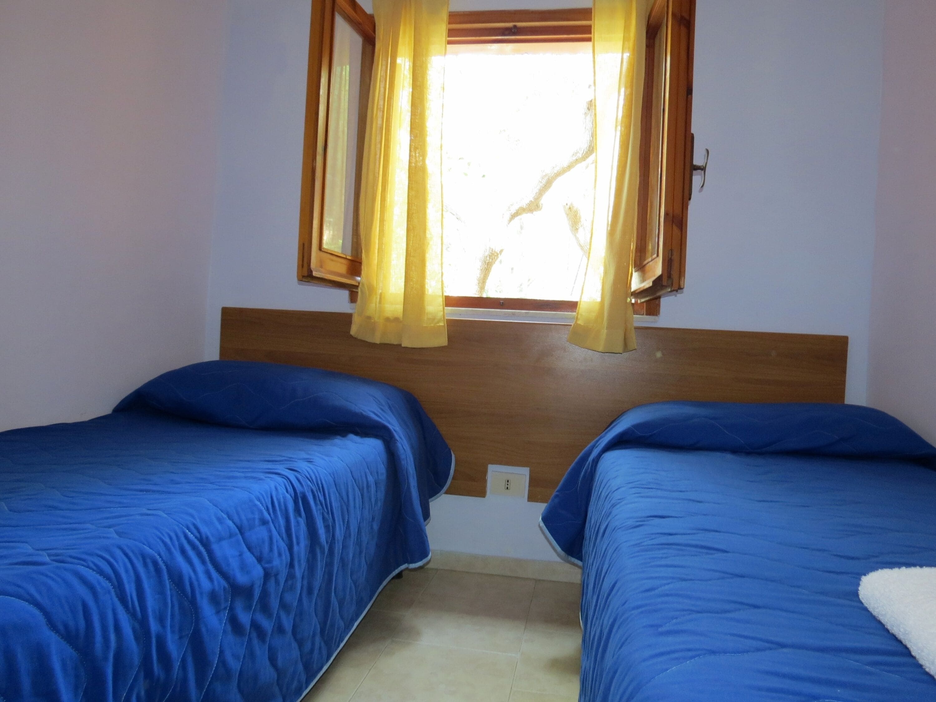 A room with two beds, a wooden headboard, a window with yellow curtains, and a light switch on the wall.