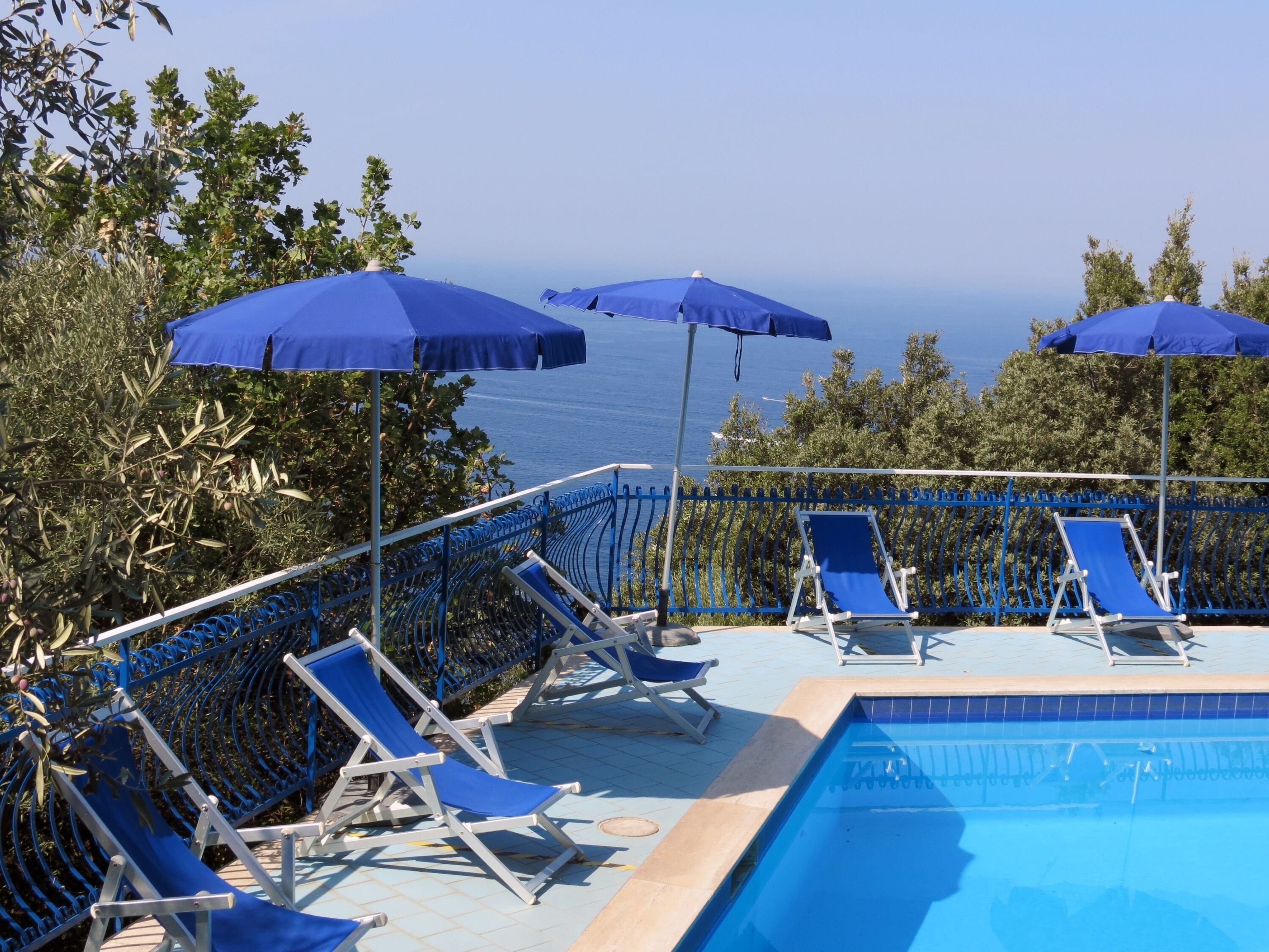 Área à beira da piscina com guarda-sóis azuis e espreguiçadeiras, com vista para um corpo d'água.