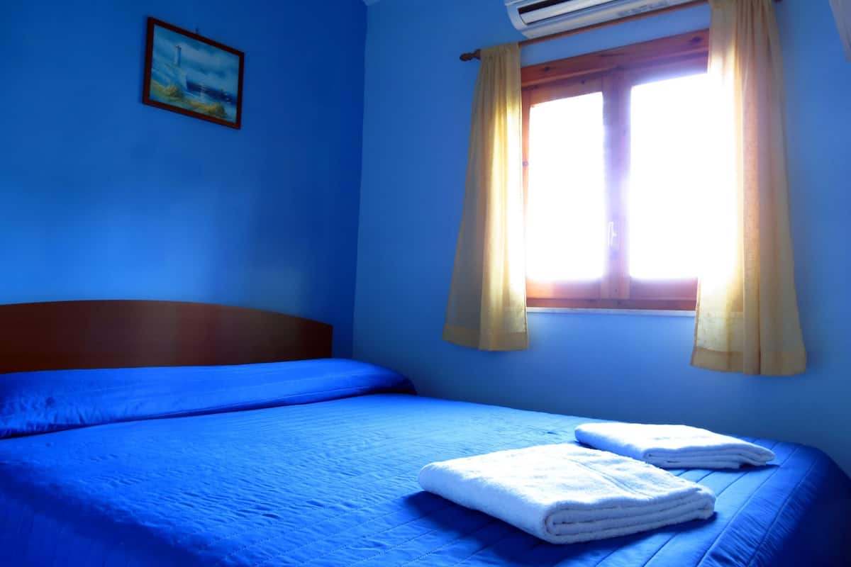 A bedroom with a bed, a window with curtains, an air conditioner, and a painting on the wall.