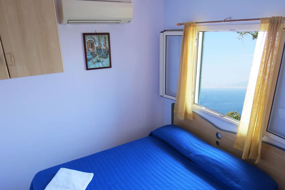 A bedroom with a bed, a window with curtains, a painting on the wall, and an air conditioning unit.