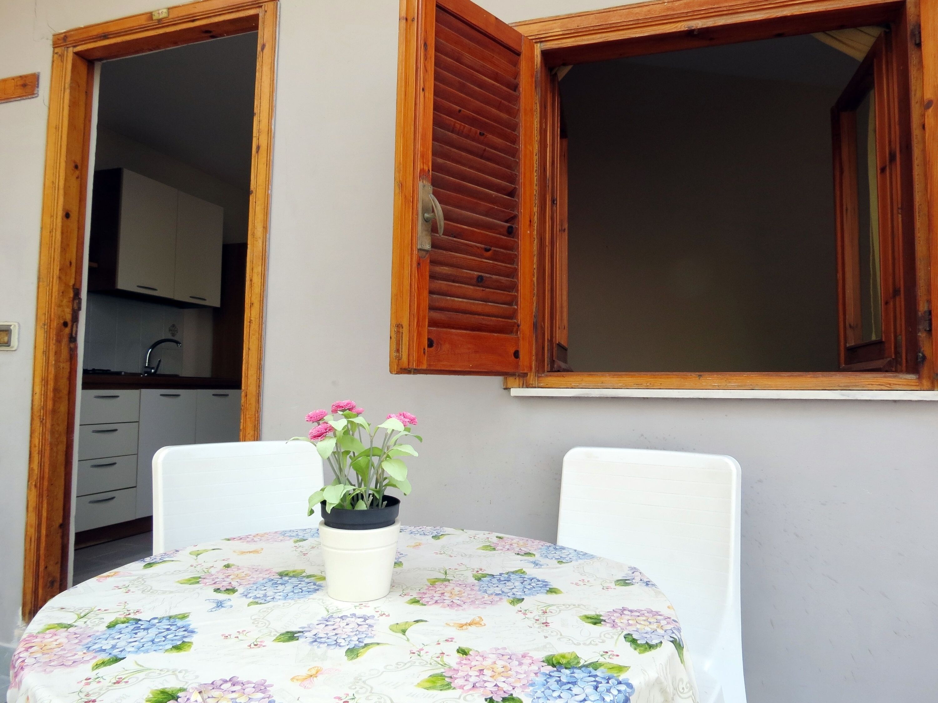 Ein kleiner runder Tisch mit einer blumigen Tischdecke, ein Blumentopf mit rosa Blüten und zwei weiße Stühle.