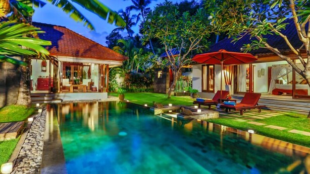 A poolside area with lounge chairs and a red umbrella, surrounded by lush greenery and a house with a tiled roof. - Villa Kaba Kaba Resort Bali (Cepaka)
