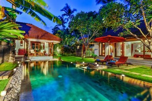 Zona de piscina con sillones de descanso y una sombrilla roja, rodeada de exuberante vegetación y una casa con tejado de tejas.