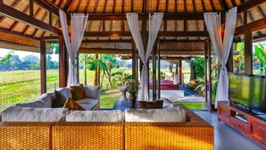 A spacious living area with a thatched roof, comfortable seating, a flat-screen TV, and a view of a lush garden. - Villa Kaba Kaba Resort Bali (Cepaka)
