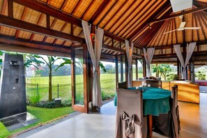 A covered outdoor dining area with a table set for two, large windows, and a view of a grassy landscape. - Villa Kaba Kaba Resort Bali (Cepaka)