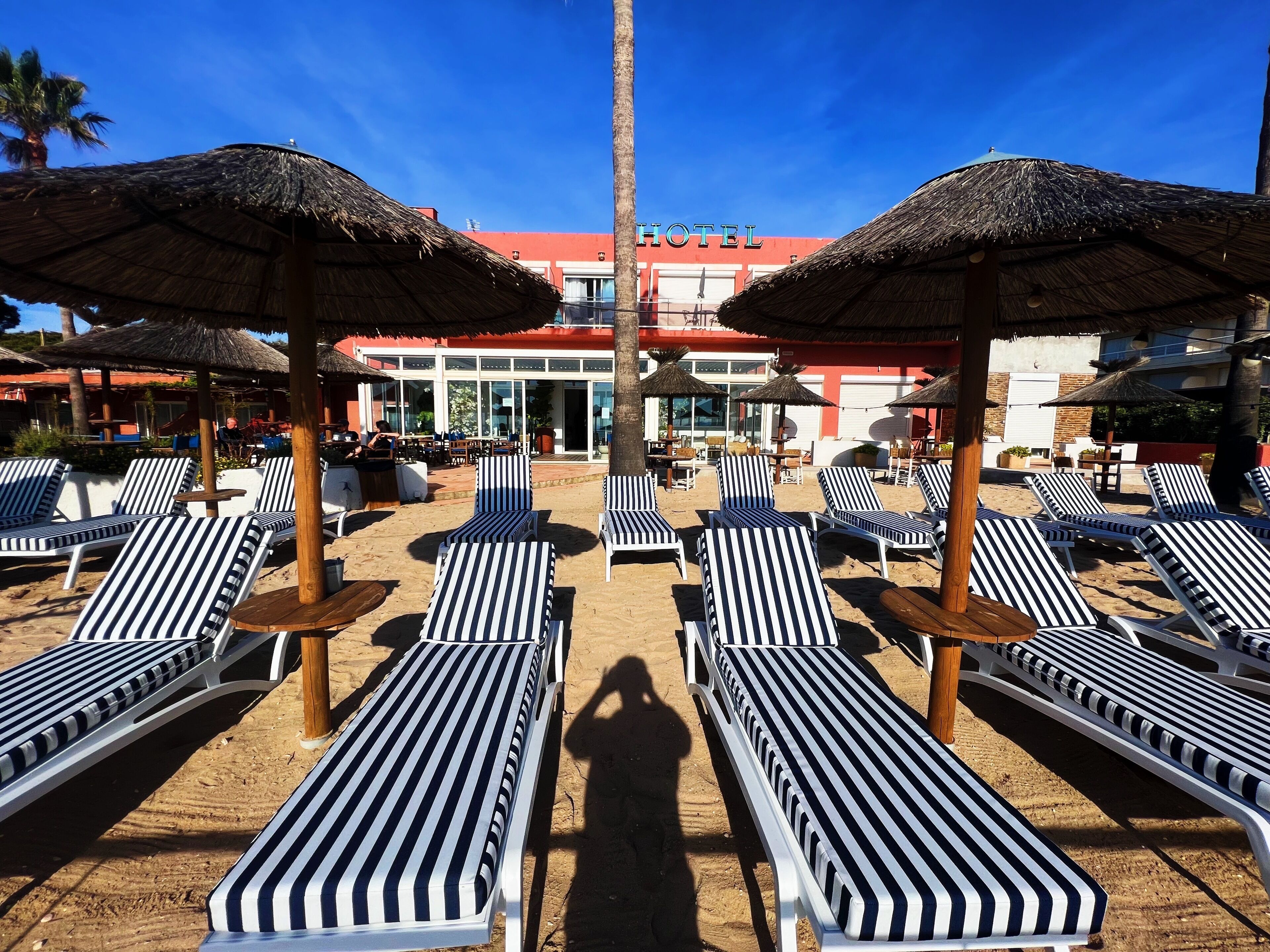 Un hotel frente a la playa con sillas de salón a rayas y sombrillas de paja, un edificio rojo al fondo y un cielo azul despejado.