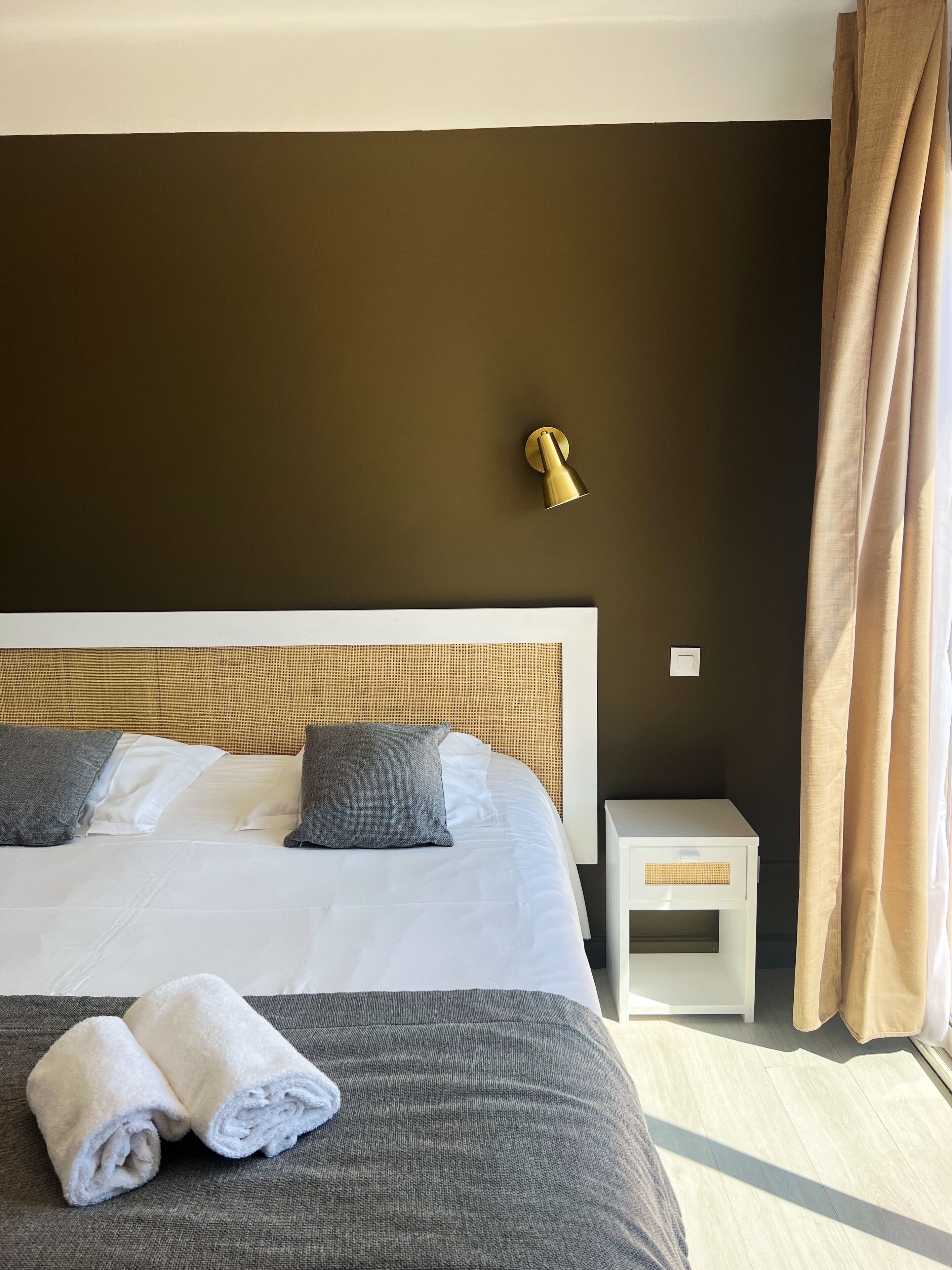 A neatly made bed with white and gray bedding, a small bedside table with a lamp, and a window with curtains.
