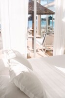 Habitación de hotel con cama, ropa de cama blanca y vistas a una zona de piscina exterior con tumbonas y sombrillas.