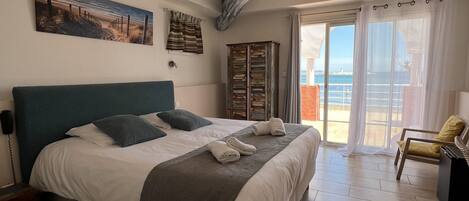 A bedroom with a bed, a painting, a wooden cabinet, and a view of the sea through a sliding glass door.