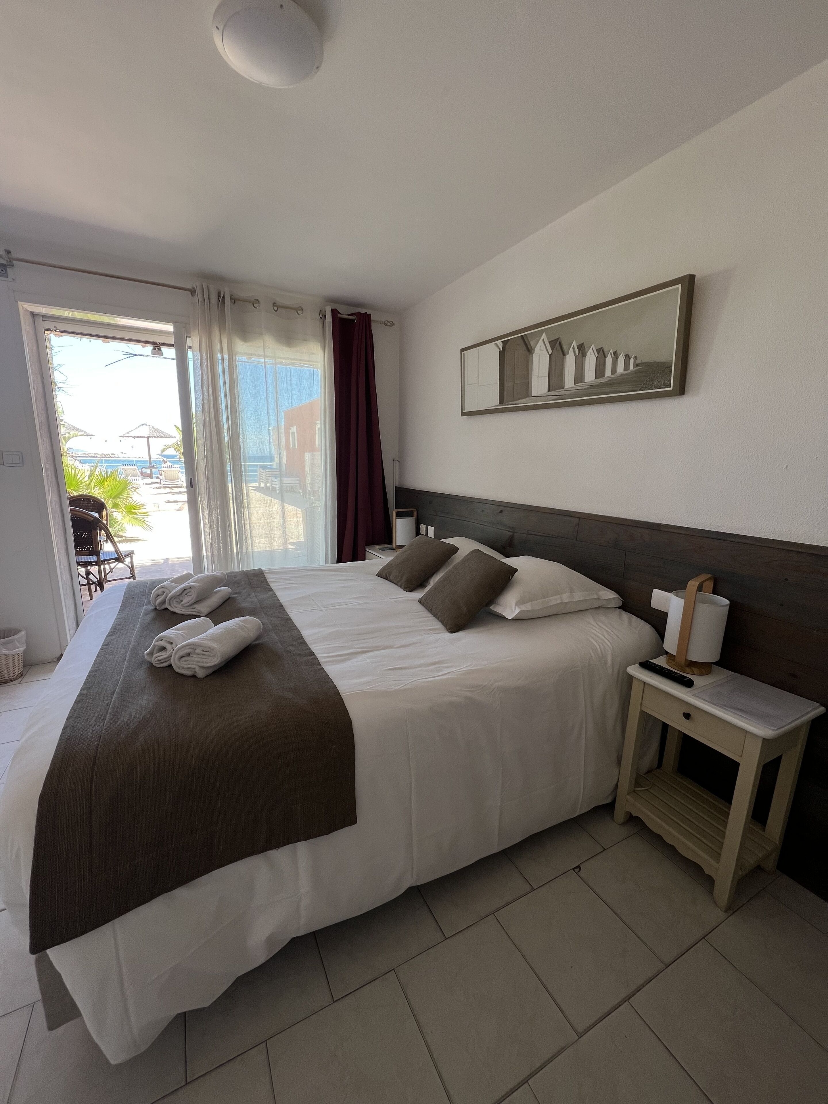 A hotel room with a large bed, a bedside table with a lamp, a mirror, and a view of the outside balcony.