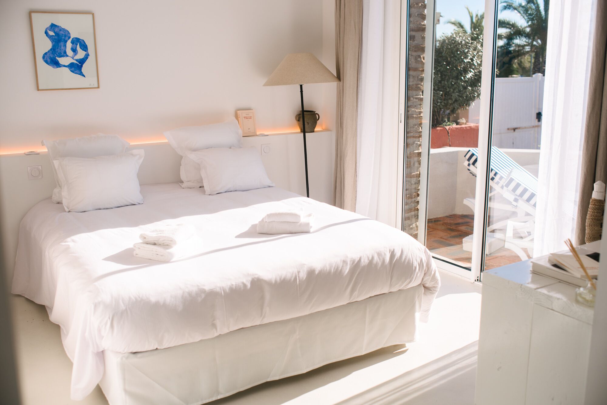 Ein Hotelzimmer mit einem großen Bett, weißer Bettwäsche, einer Nachttischlampe, einem gerahmten Kunstwerk und Blick ins Freie.