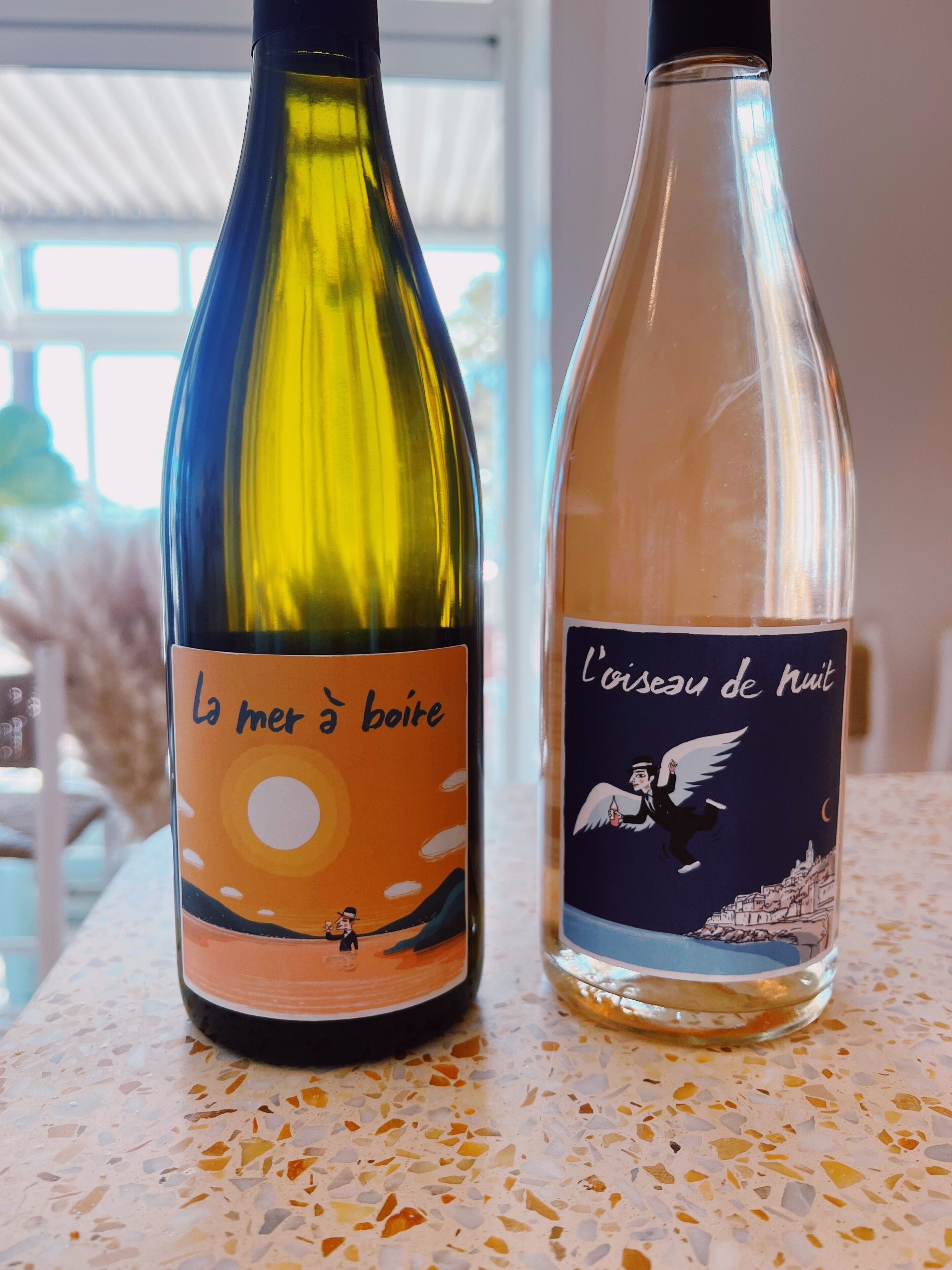 Two wine bottles on a terrazzo table with a view of a building and trees in the background.