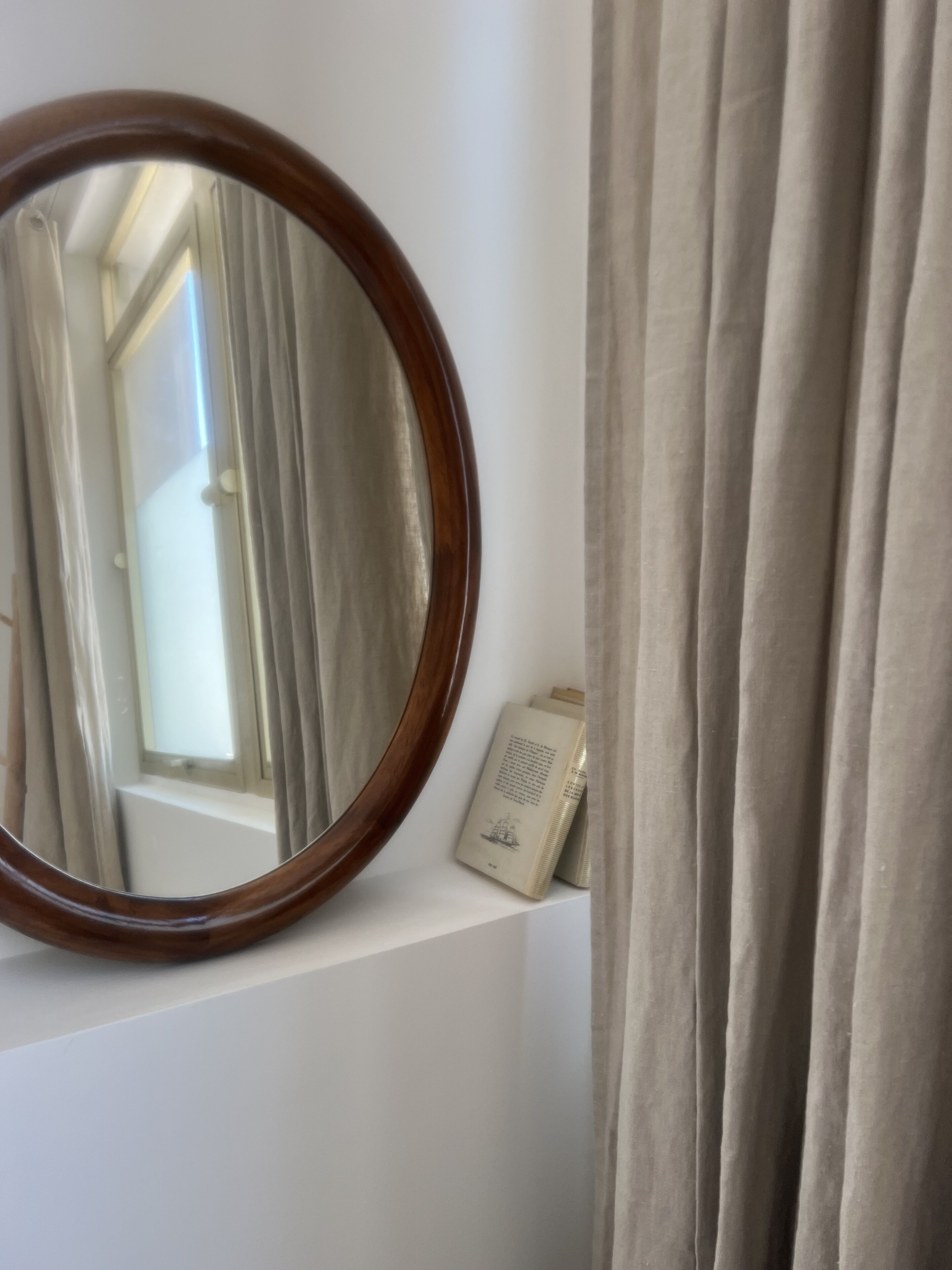 A round mirror with a wooden frame on a white wall, a small book, and a curtain partially drawn to the side.