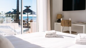 Habitación de hotel con un ventanal grande, una cama con sábanas blancas, una mesita de noche pequeña con una lámpara y vistas a la playa y sombrillas.