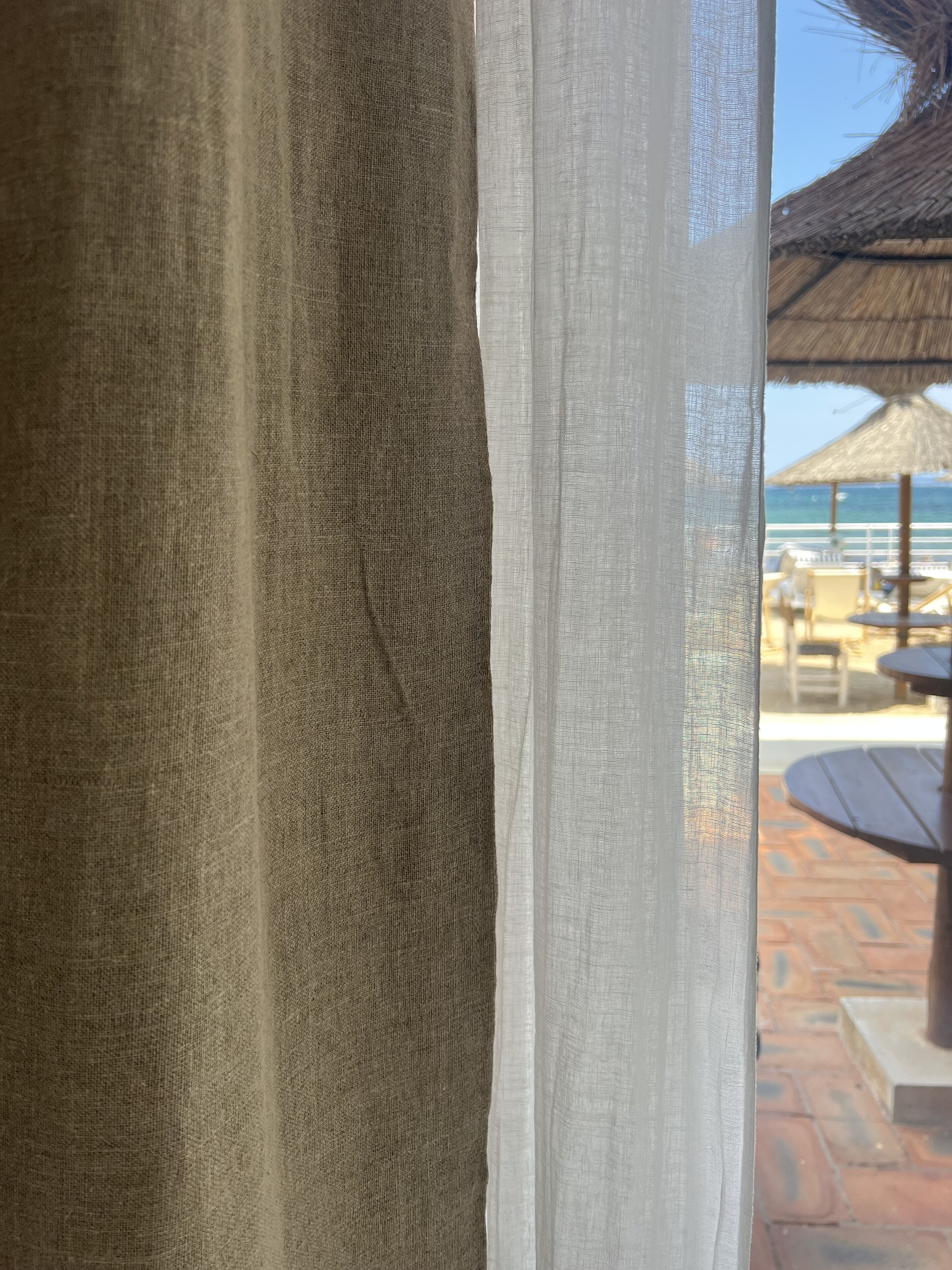 A view through a window with a textured curtain, a thatched umbrella, and outdoor furniture.