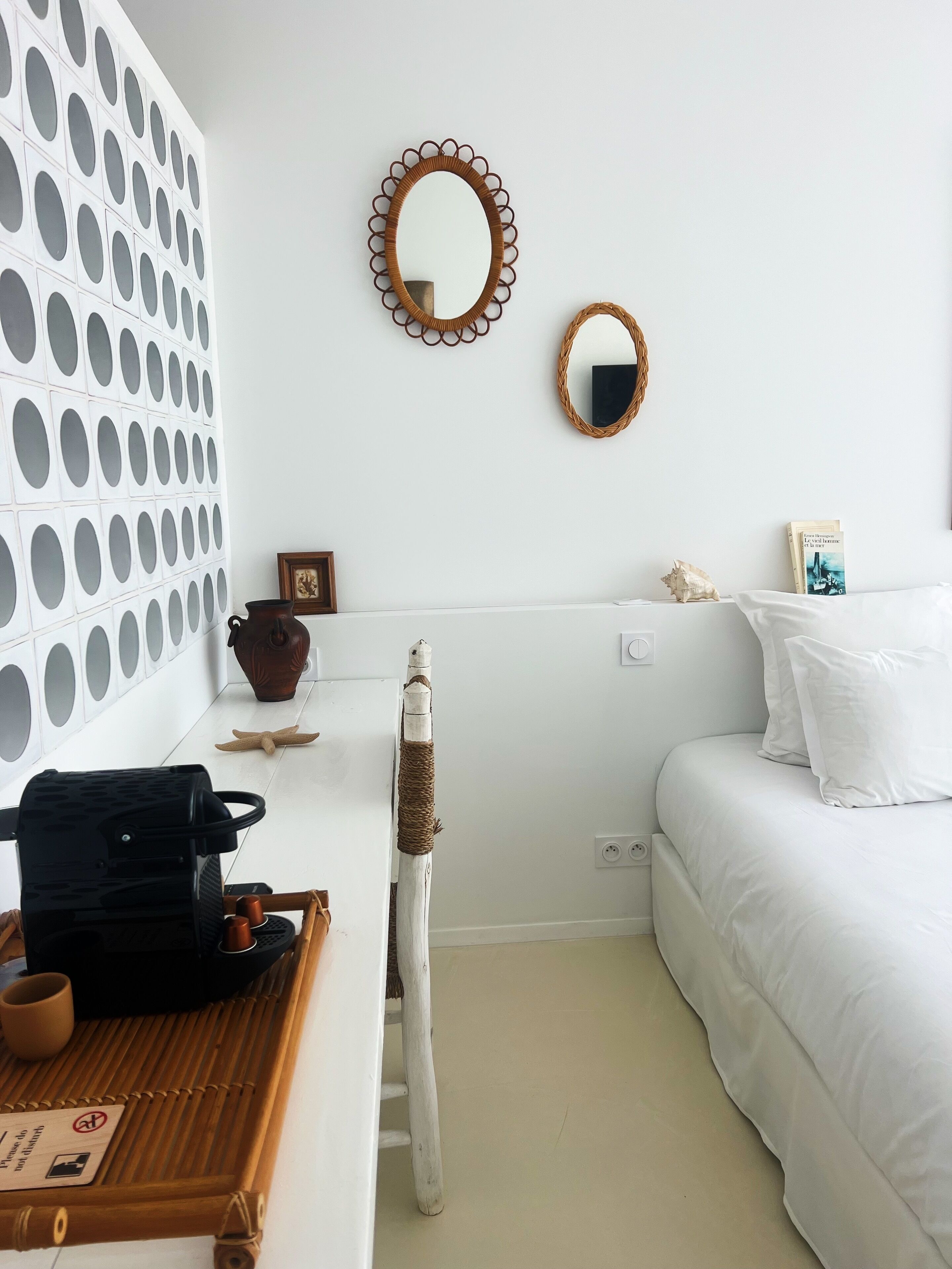 A bedroom with a white bed, a small bedside table with a book, a mirror, and a wall with circular patterns.