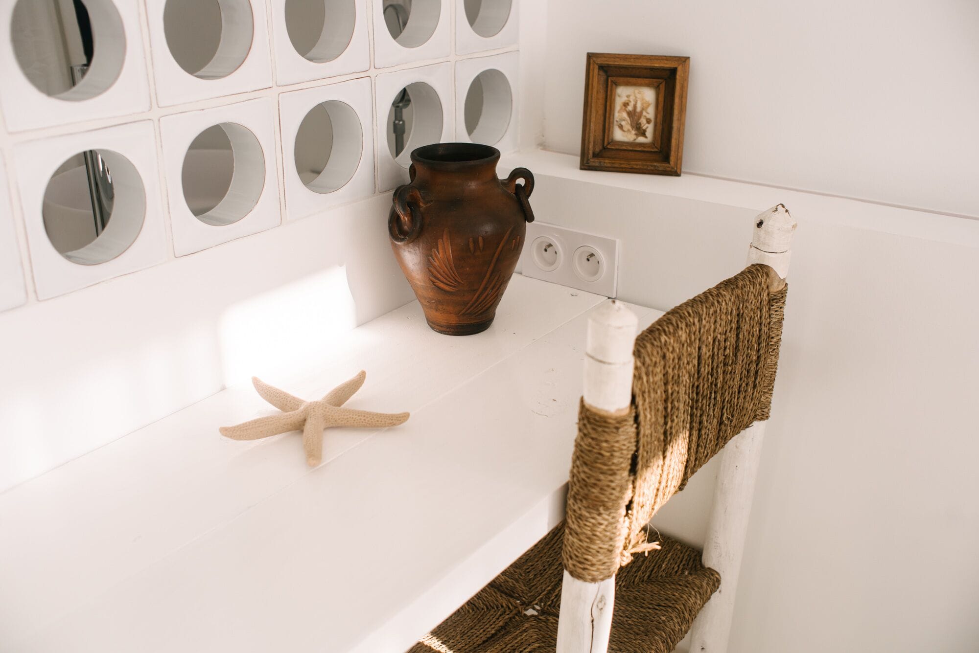 Un baño con un lavabo blanco, una decoración de estrella de mar, un jarrón de cerámica marrón y un cuadro en la pared.