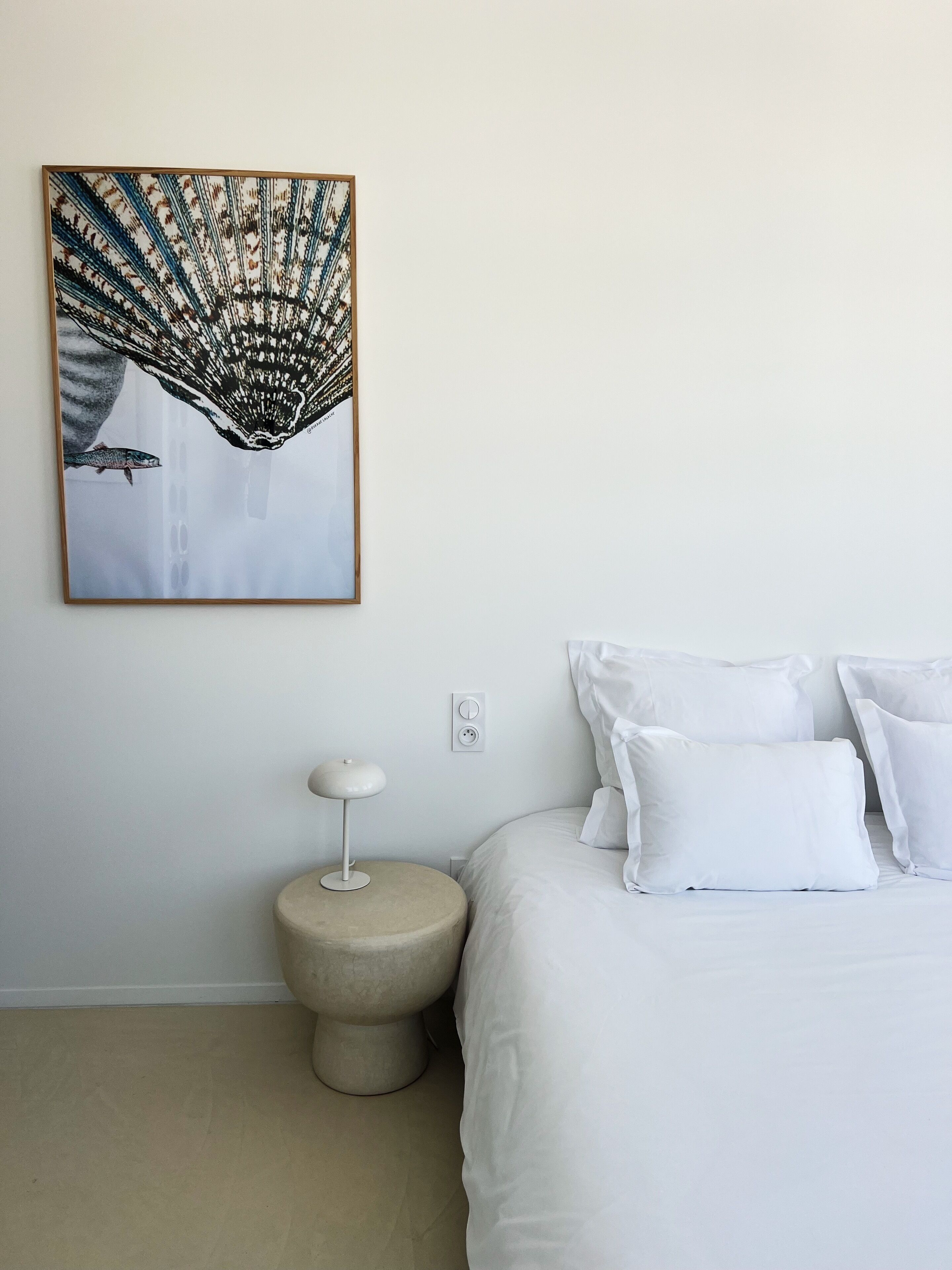 A bedroom with a bed, white pillows, a round nightstand, a modern lamp, and a wall-mounted abstract painting.