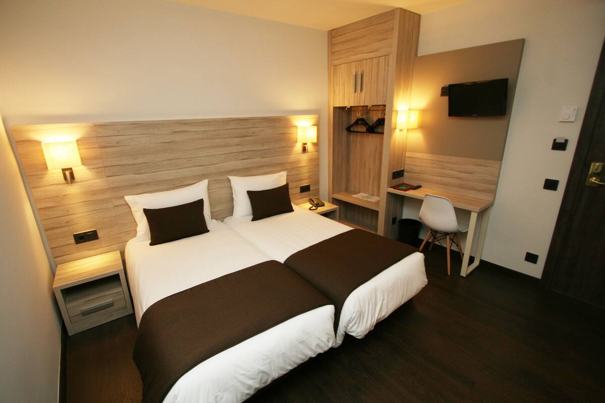 Ein Doppelbett mit zwei Kissen, ein Nachttisch, ein Schreibtisch mit Stuhl und ein an der Wand befestigter Fernseher.