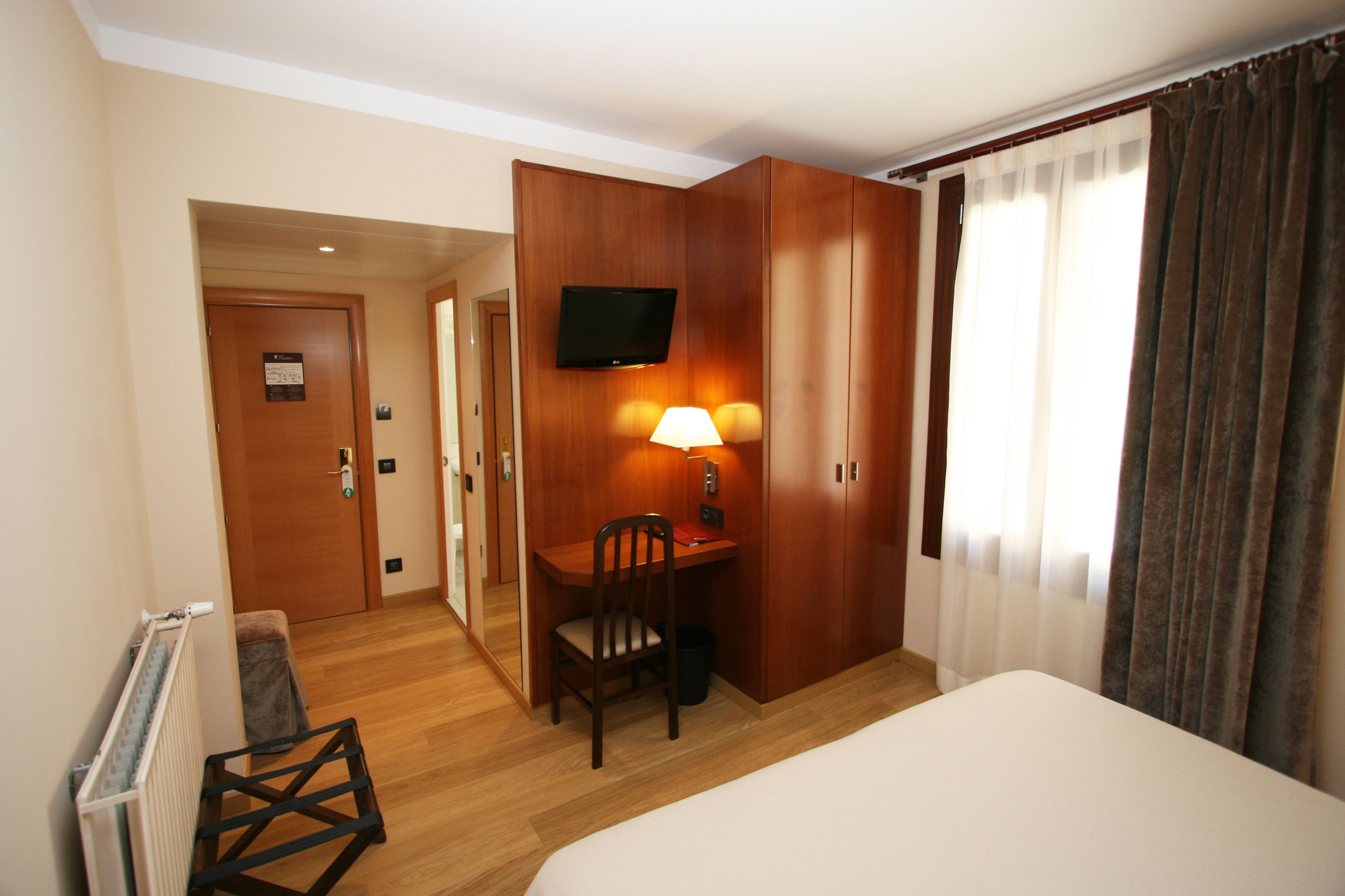 Habitació d'hotel amb un llit, una taula de treball amb una cadira, un televisor penjat a la paret i una finestra amb cortines.