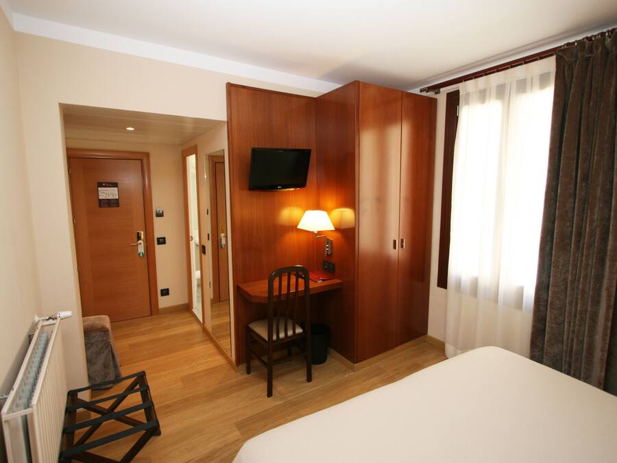 Habitació d'hotel amb un llit, una taula de treball amb una cadira, un televisor penjat a la paret i una finestra amb cortines.