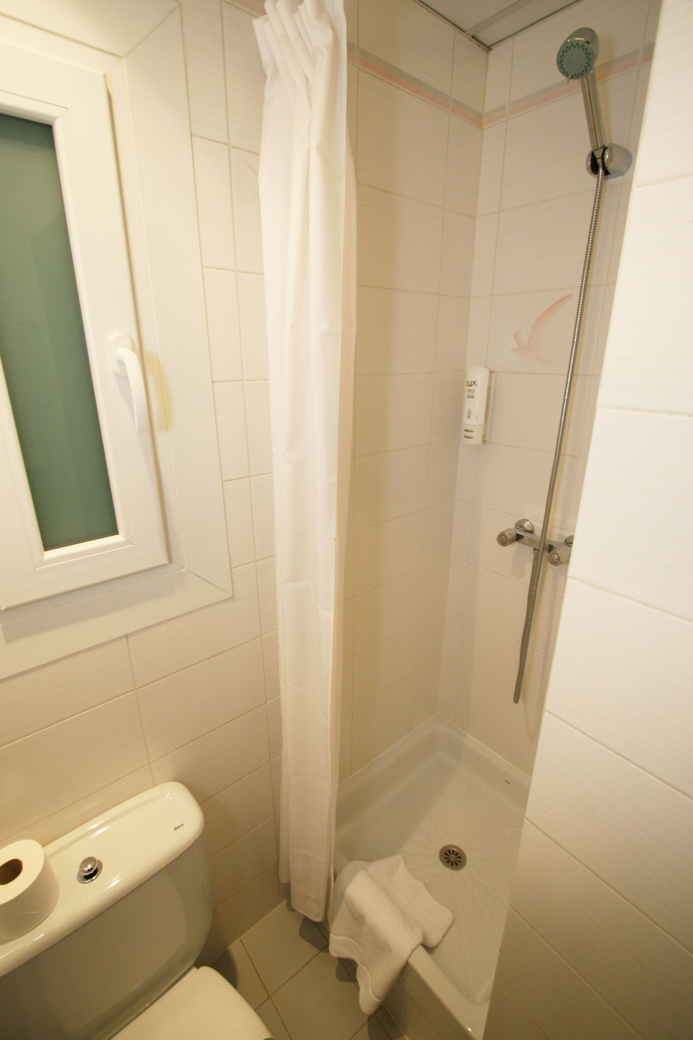 Um espaço de chuveiro pequeno e fechado, com porta de vidro, uma toalha branca pendurada na parede e um vaso sanitário com dispensador de papel.