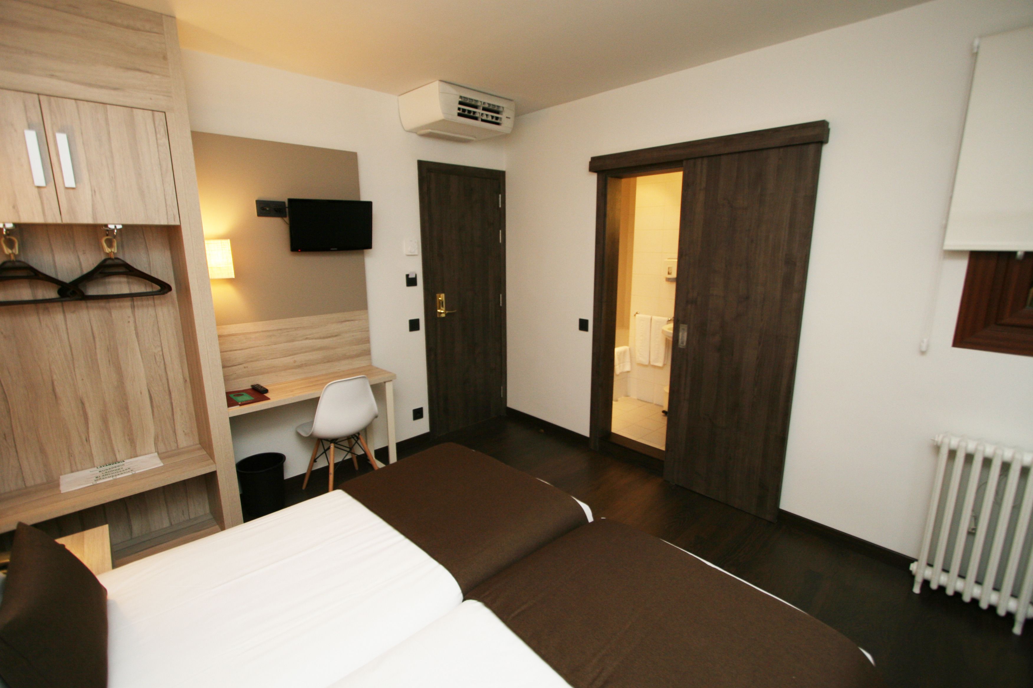 Habitación de hotel con cama, escritorio, silla, armario, televisor y puerta corredera que da al baño.