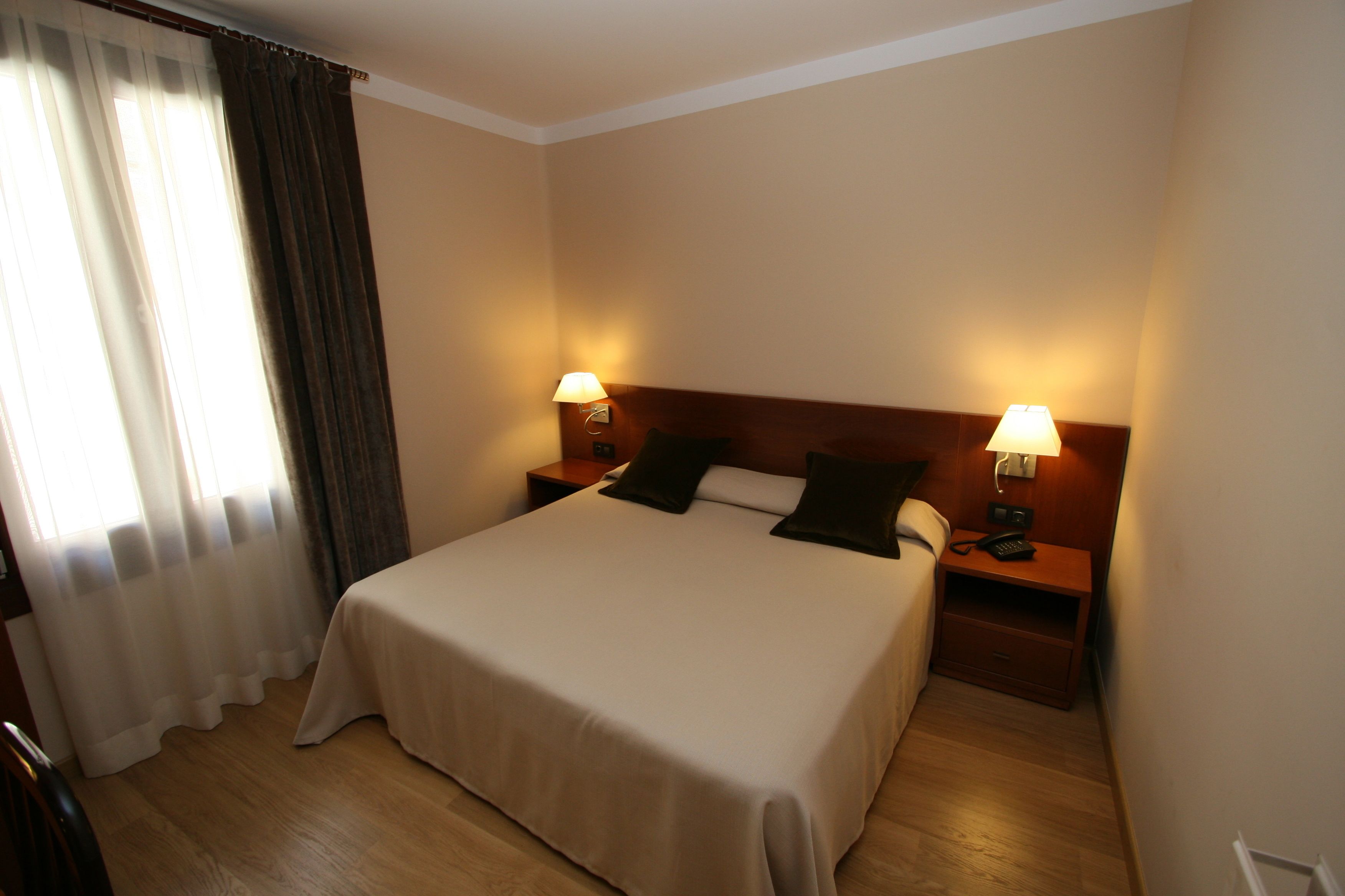 A hotel room with a large bed, two bedside tables with lamps, a wooden headboard, and a window with curtains.